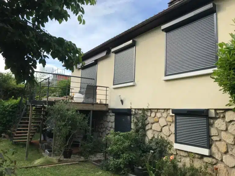 Maison avec volets roulants gris à l'étage et au rez-de-chaussée, jardin au premier plan