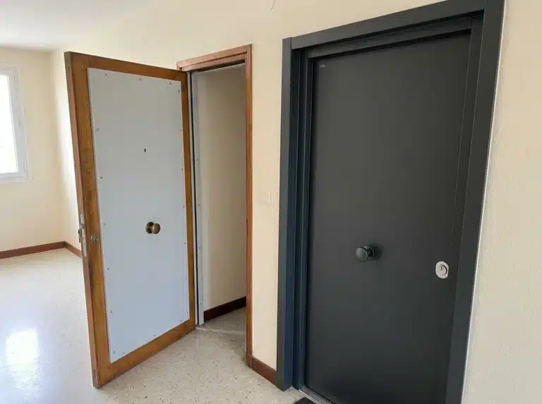 Sur un palier, une porte en bois entrouverte montre son renfort en acier, près d’une porte blindée et de son bâti.