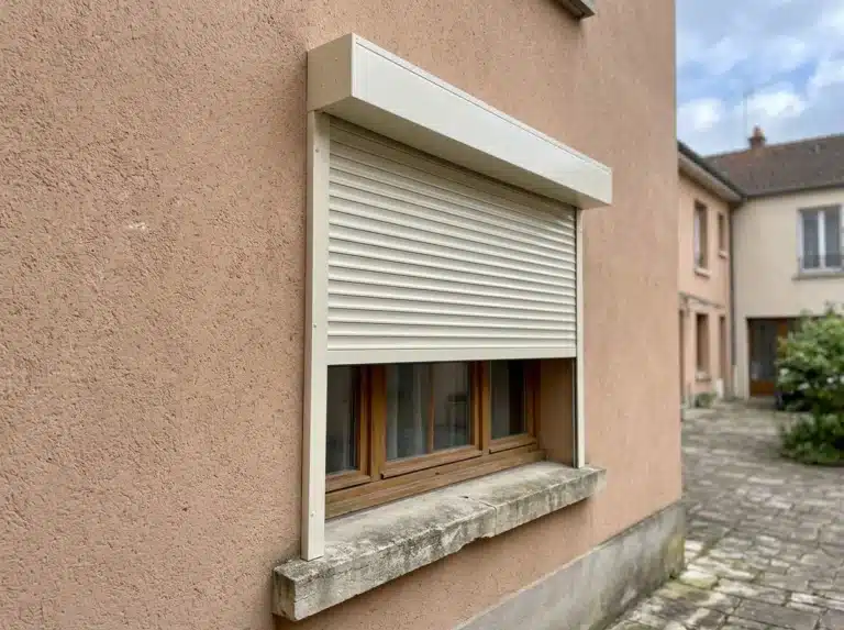 Volet roulant ivoire à mi-hauteur sur une fenêtre, avec coffre extérieur et coulisses sur une façade terracotta.