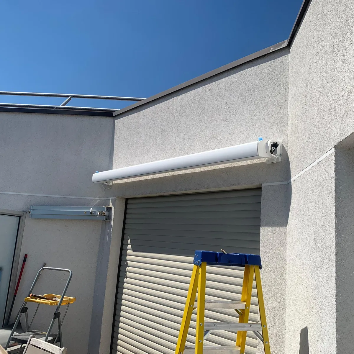 Coffre de store banne blanc fixé sur une façade au-dessus d'une baie, escabeau jaune au pied