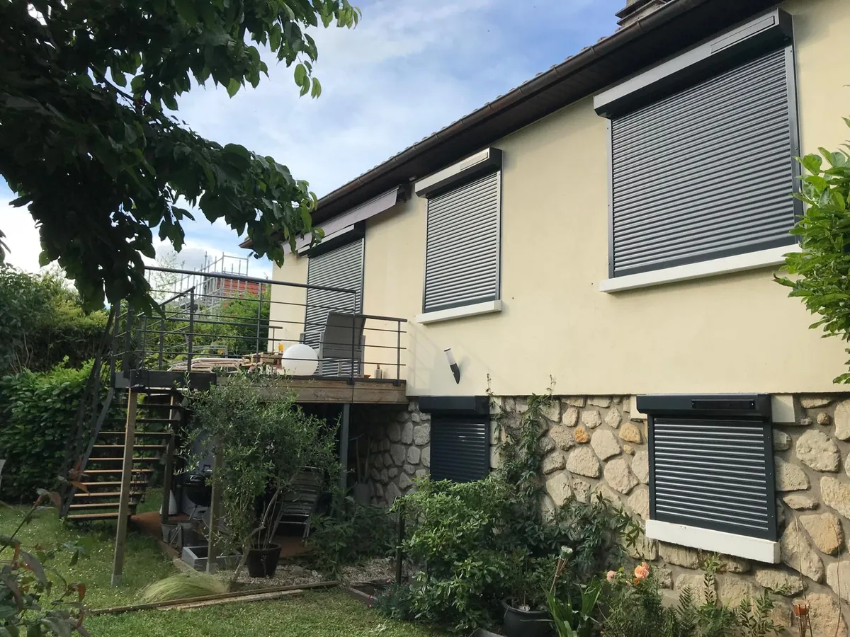 Maison avec volets roulants gris, escalier extérieur et jardin arboré